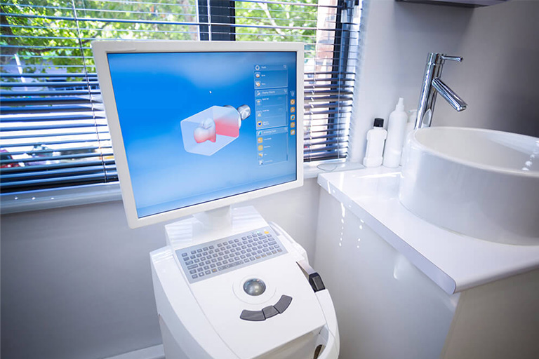 A dental CAD/CAM (Computer-Aided Design and Computer-Aided Manufacturing) system in a dental office. The setup includes a computer with a monitor displaying a 3D model of a dental restoration, and a milling machine used to fabricate the dental restoration based on the digital design.