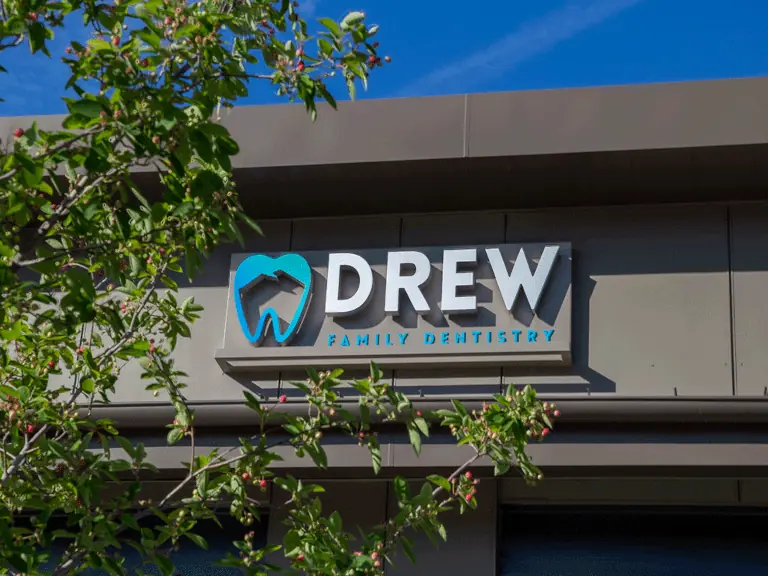 Exterior sign for Drew Family Dentistry mounted on a gray building. The sign features a blue and white logo of a stylized tooth with a house inside it, accompanied by the text 'DREW' in bold white letters and 'FAMILY DENTISTRY' in smaller blue letters. Green leaves partially frame the sign, with a clear blue sky in the background.