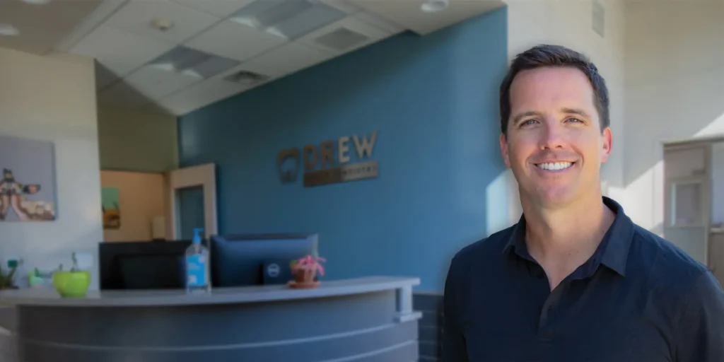 The reception area of Drew Family Dentistry with a curved dark desk featuring a computer monitor, hand sanitizer, and a potted plant on top. Behind the desk, Dr. Drew's headshot in black attire is visible, standing in front of a blue wall displaying the 'Drew Family Dentistry' logo with a tooth icon.
